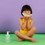 A young girl in a yellow swimsuit sits cross-legged, holding a paper honeydew melon cutout in front of her face, next to Dabble & Dollop honeydew gel.
