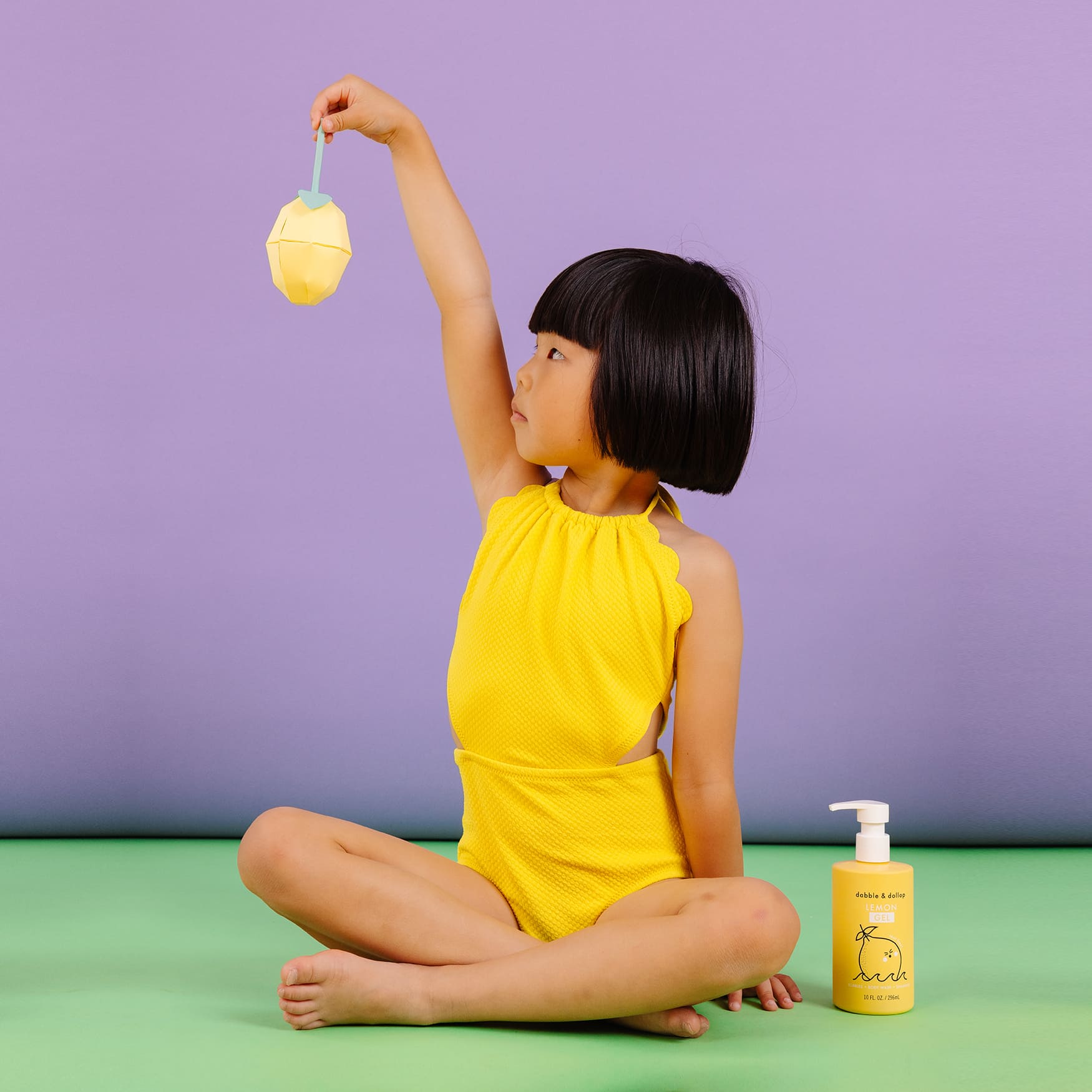 A young girl in a yellow swimsuit holds up a paper lemon ornament next to Debble & Dollop lemon gel soap.
