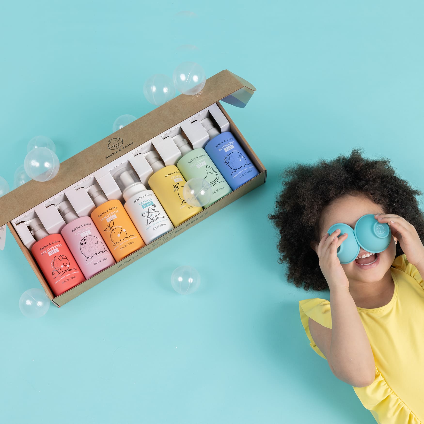 A happy girl wearing a yellow swimsuit looks through blue Dabble & Dollop bubble bath containers next to a gift box of rainbow-colored kids' body wash and shampoo.
