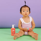 A baby sits beside Dabble & Dollop strawberry gel, holding a strawberry-shaped toy.
