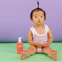 A baby sits beside Dabble & Dollop strawberry gel, holding a strawberry-shaped toy.
