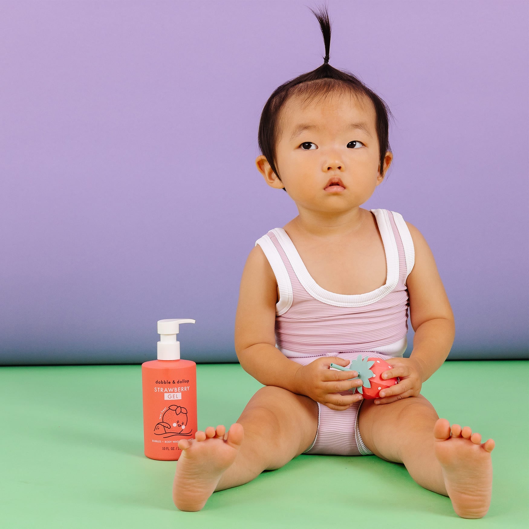 A baby sits beside Dabble & Dollop strawberry gel, holding a strawberry-shaped toy.
