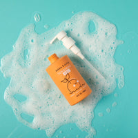 An orange bottle of Dabble & Dollop Tangerine Gel body wash and shampoo sits amid frothy bubbles on a teal background.
