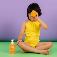 A young girl in a yellow swimsuit playfully peeks through a tangerine toy, advertising Dabble & Dollop tangerine kids' shampoo.
