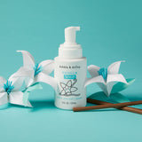 Dabble & Dollop Vanilla Whip bubbles, body wash, and shampoo sit beside paper flowers on a teal background.
