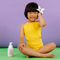 A smiling Asian girl in a yellow swimsuit sits cross-legged with a flower in her hair next to a Dabble & Dollop Vanilla Whip children's body wash and shampoo.
