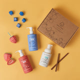 Dabble & Dollop's mixable kids' bath products, including strawberry gel, blueberry gel, and vanilla whip body wash and shampoo, are playfully styled with paper fruit on a yellow background.
