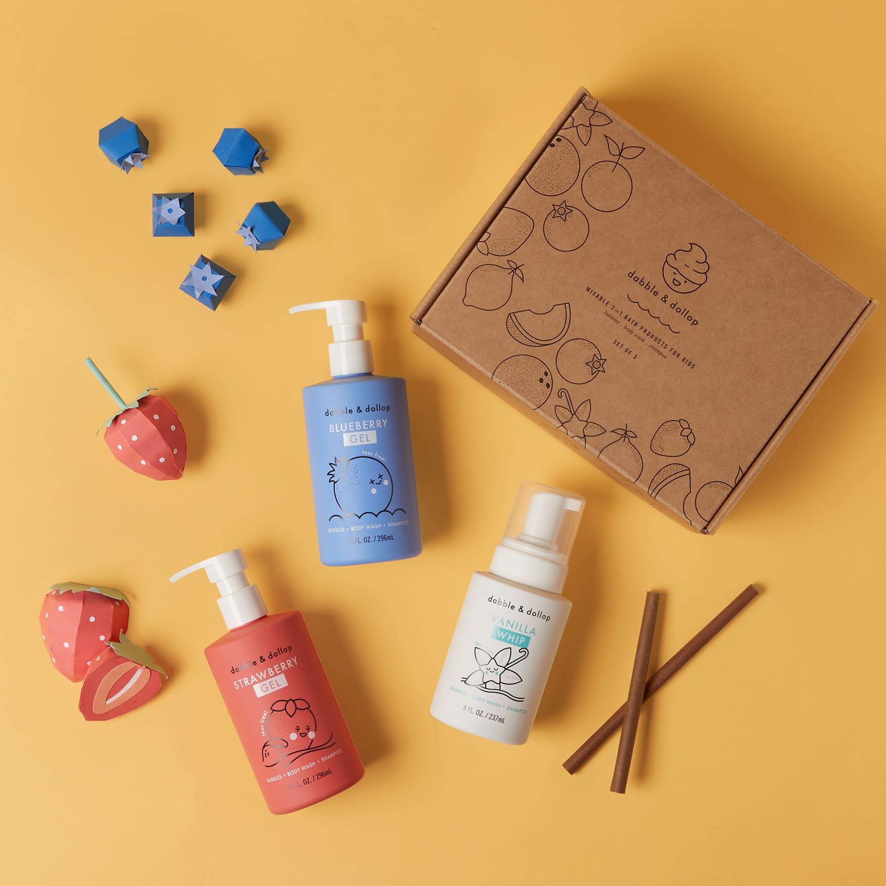 Dabble & Dollop's mixable kids' bath products, including strawberry gel, blueberry gel, and vanilla whip body wash and shampoo, are playfully styled with paper fruit on a yellow background.

