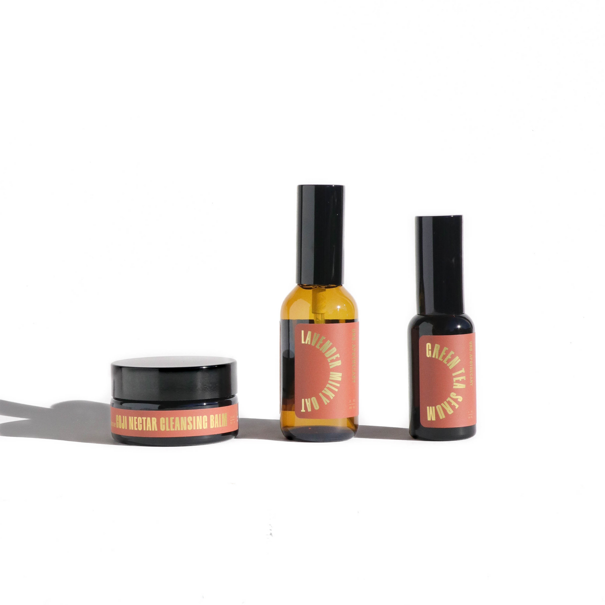 Close-up shows three skincare products: Goji Nectar Cleansing Balm, Lavender Milky Oat spray, and Green Tea Serum on a white background.
