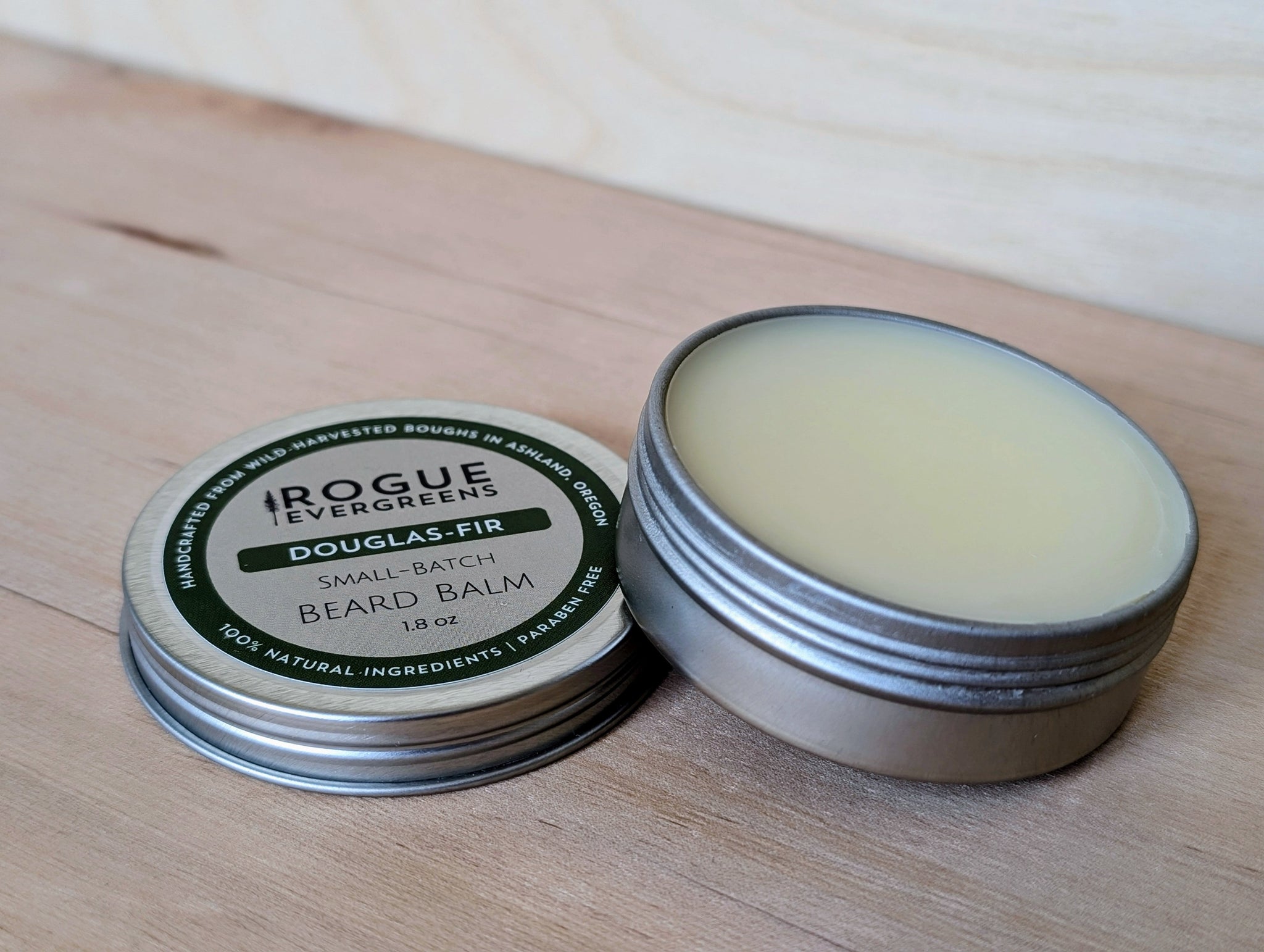 Two tins of Rogue Evergreens Douglas-Fir beard balm sit on a wooden surface.
