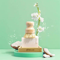 Green Llama promotes eco-friendly dishwashing with a natural bristle brush resting on coconut soap beside white flowers.
