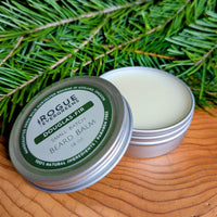 A close-up shows two tins of Rogue Evergreens Douglas Fir beard balm, one open, against a background of fir boughs.
