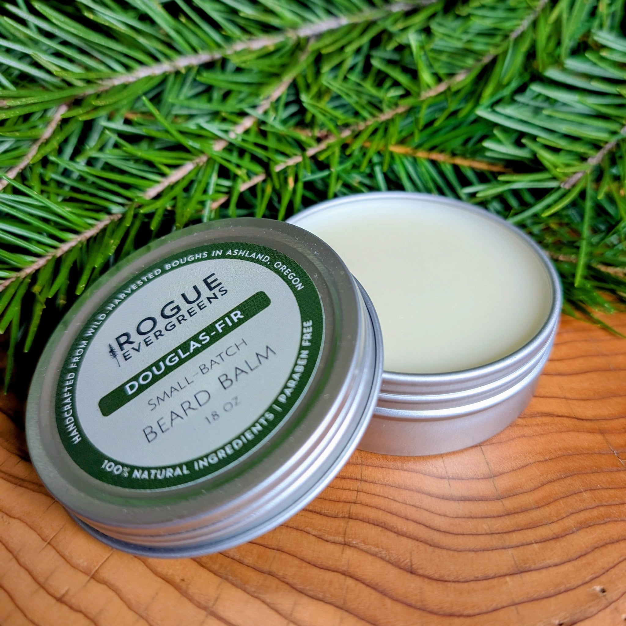 A close-up shows two tins of Rogue Evergreens Douglas Fir beard balm, one open, against a background of fir boughs.
