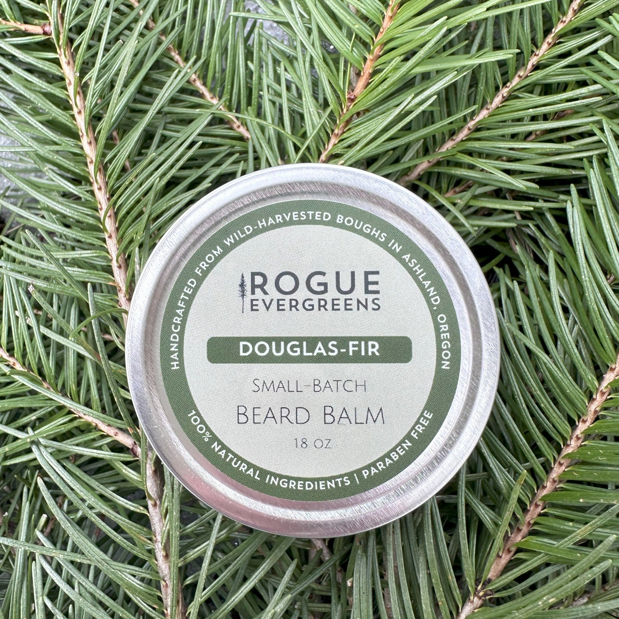 Rogue Evergreens Douglas-Fir beard balm rests on fresh evergreen boughs.
