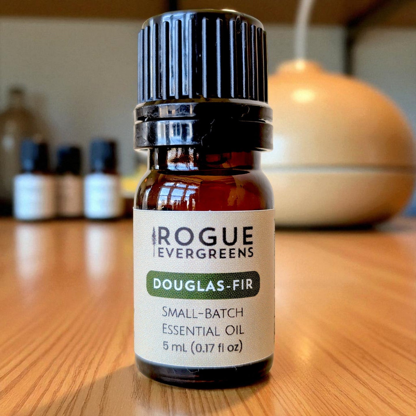 A bottle of Rogue Evergreens Douglas-Fir essential oil sits on a wooden table near a diffuser.
