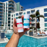Hand holds two Breathe Naturals deodorant sticks, Watermelon Shaved Ice and Cooling Cocomint, poolside at a resort.
