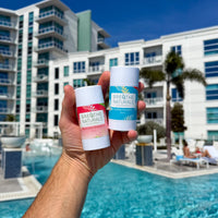 Hand holds two Breathe Naturals deodorant sticks, Watermelon Shaved Ice and Cooling Cocomint, poolside at a resort.

