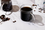 Two glass coffee mugs hold freshly brewed dark roast coffee next to scattered beans and a bag of gourmet Colombian coffee.
