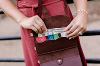 A woman's hands place a twenty-dollar bill and credit cards into a brown leather Urban Southern crossbody wallet bag.
