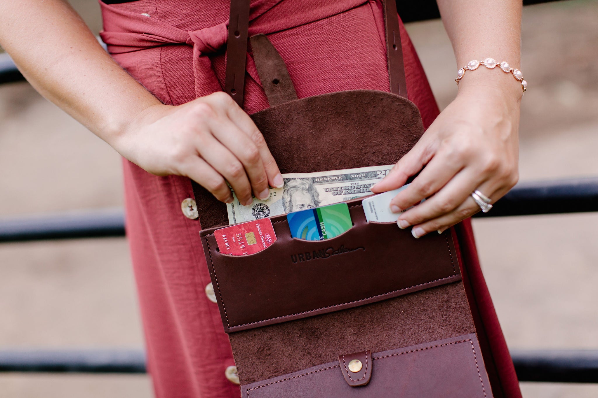 A woman's hands place a twenty-dollar bill and credit cards into a brown leather Urban Southern crossbody wallet bag.
