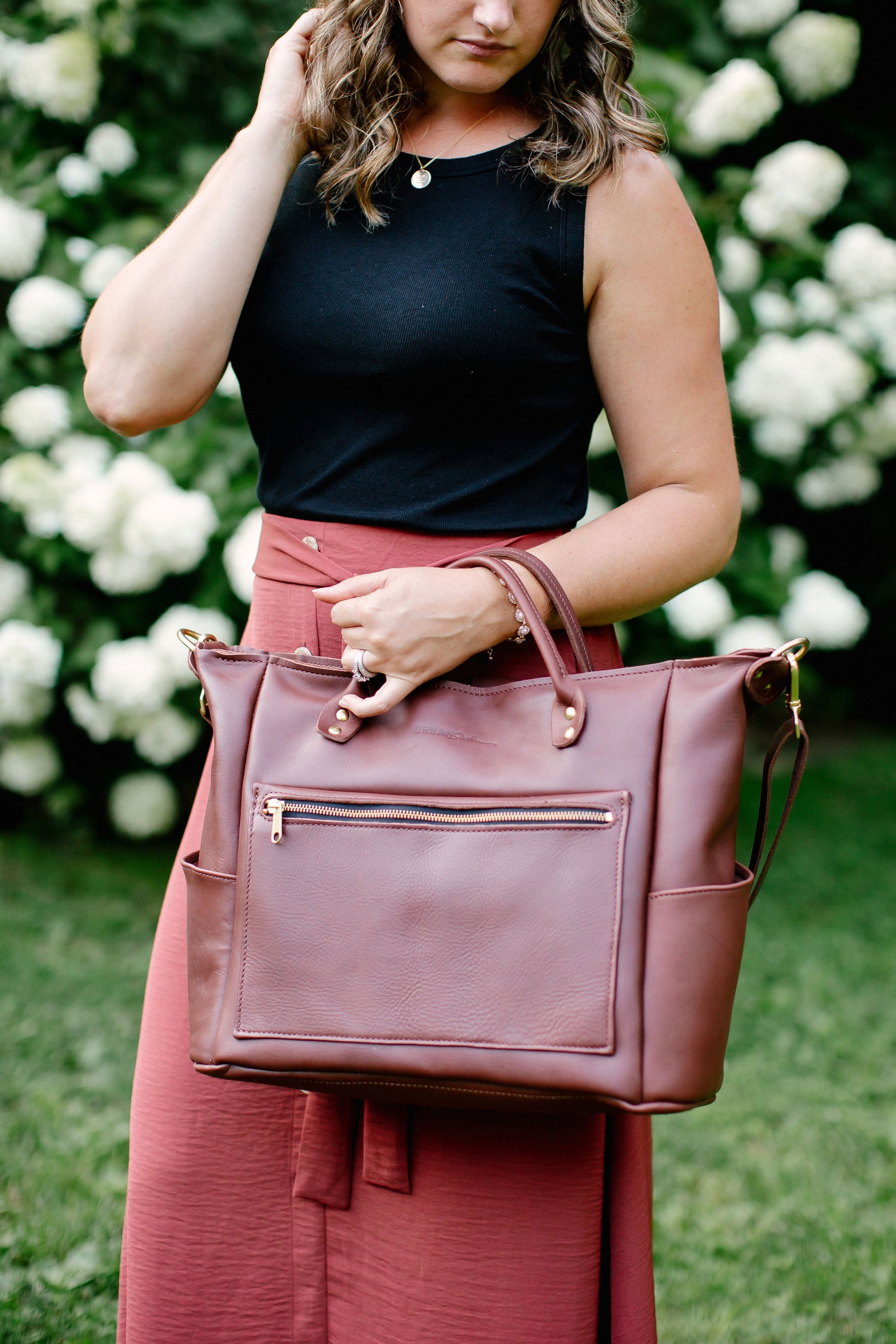 A woman models a stylish burgundy leather tote bag with a front zipper pocket, standing against a backdrop of white hydrangeas.
