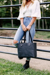 A woman wearing ripped jeans and combat boots holds a black leather tote bag while leaning against a metal fence.
