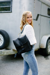 A blonde woman models a stylish black leather backpack with a gold ring detail in front of a horse trailer.
