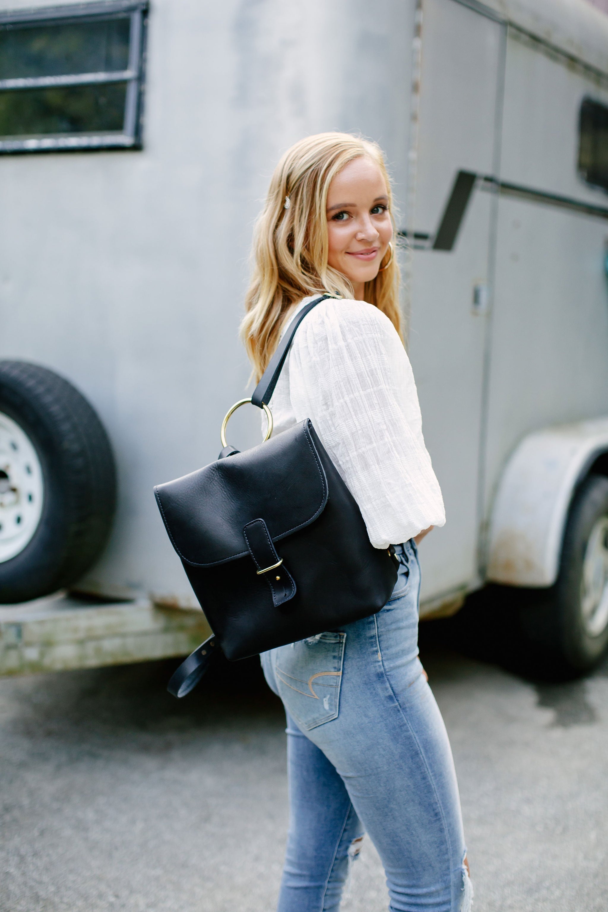 A blonde woman models a stylish black leather backpack with a gold ring detail in front of a horse trailer.
