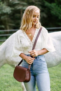 A blonde woman wearing a white blouse and blue jeans holds a brown leather saddle bag, standing next to a white horse in a grassy field.
