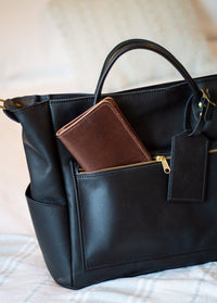 A brown leather wallet sits inside the zippered pocket of a stylish black leather tote bag.
