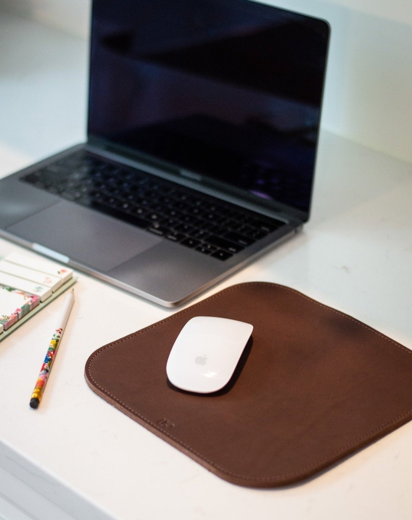 A laptop sits on a white desk next to a leather mousepad with a white Apple mouse and a colorful pen and notepad.
