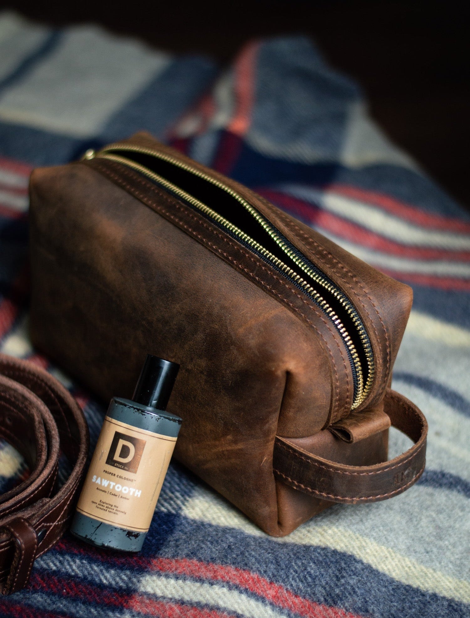 Showcasing a rustic leather dopp kit next to a bottle of "Sawtooth" men's cologne and a leather belt on a striped blanket.
