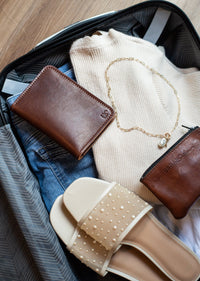 An open suitcase reveals carefully packed travel essentials: a sweater, jeans, elegant pearl-studded sandals, a leather wallet, a coin purse, and a delicate gold necklace.
