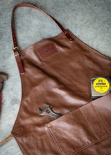 A brown leather apron rests on a concrete surface, featuring a hammer, adjustable wrench, and Stanley Powerlock tape measure in its pockets, showcasing craftsmanship and utility.

