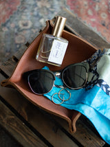 A leather tray displays sunglasses, gold hoop earrings, "Femme Fatale" perfume, and a silk scarf.
