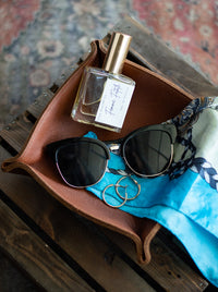 A leather tray displays sunglasses, gold hoop earrings, "Femme Fatale" perfume, and a silk scarf.
