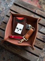 Leather valet tray holds a card wallet, sugar skull Zippo lighter, keys, and a Swiss Army knife on a rustic wood surface.
