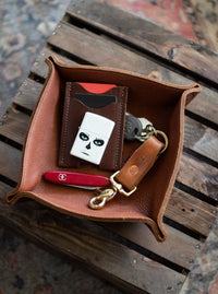 Leather valet tray holds a card wallet, sugar skull Zippo lighter, keys, and a Swiss Army knife on a rustic wood surface.
