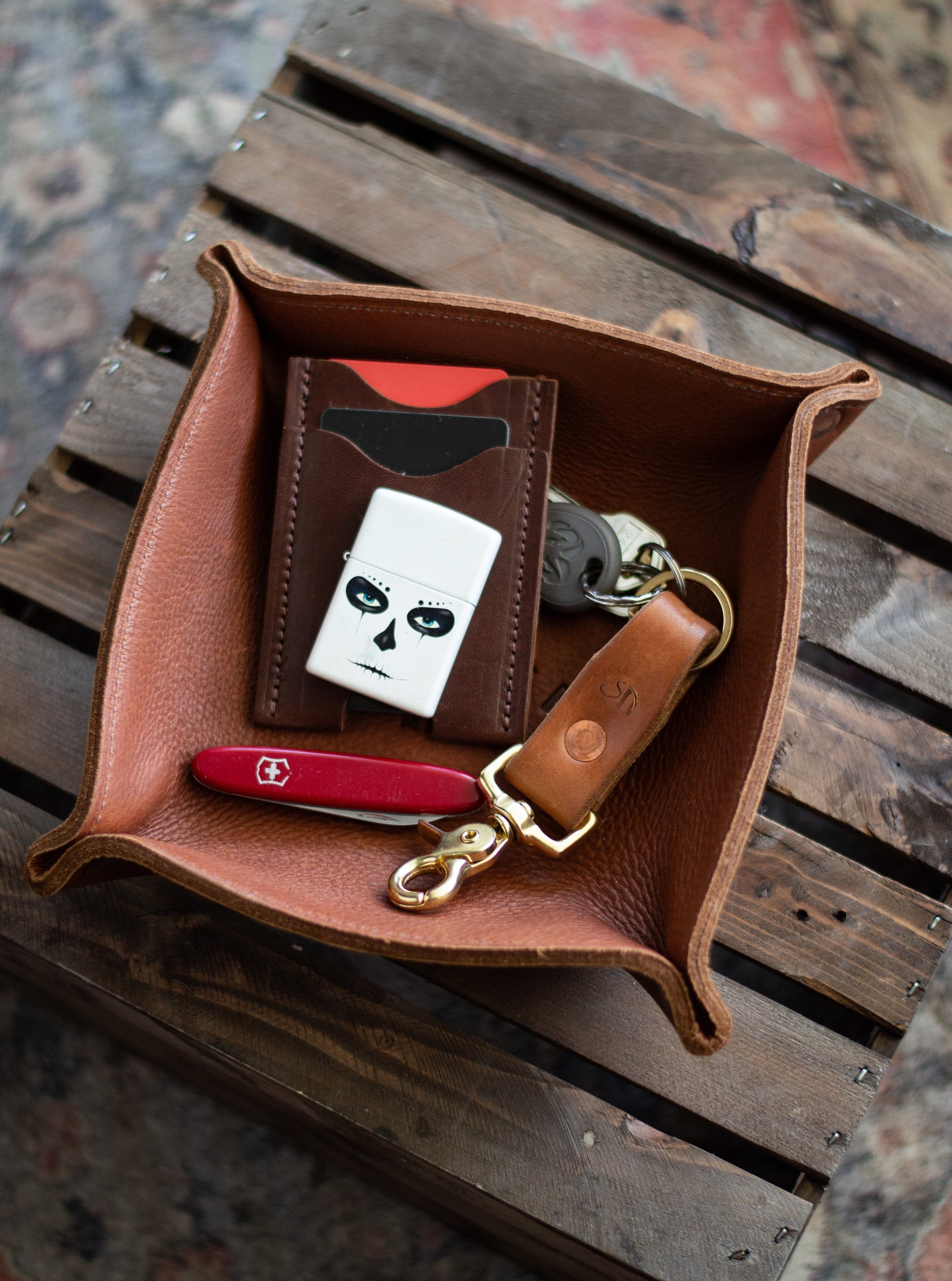 Leather valet tray holds a card wallet, sugar skull Zippo lighter, keys, and a Swiss Army knife on a rustic wood surface.
