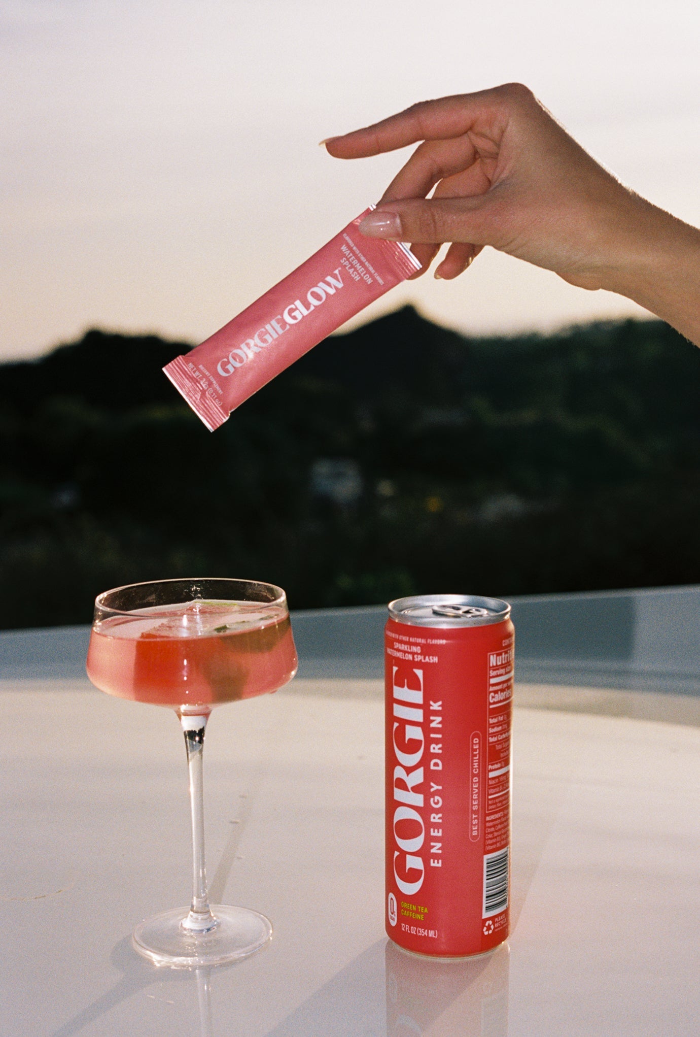 A hand holds a Gorgie Glow supplement packet above a Gorgie energy drink can and cocktail glass filled with a refreshing watermelon splash beverage.
