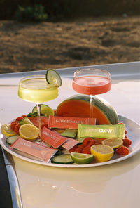 Two cocktail glasses flank a watermelon half on a platter featuring Gorgie Glow drink mix packets, lemons, limes, cucumbers, and raspberries.
