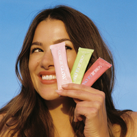 A smiling woman holds up three colorful packets of GORGIE GLOW beauty lemonade, a wellness supplement.
