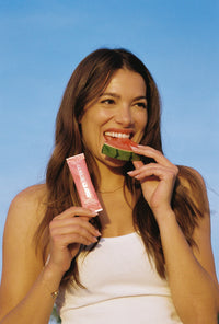 A smiling young woman in a white tank top bites into a slice of watermelon while holding a Gorgie Glow Watermelon Splash supplement packet.
