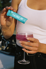 A woman prepares a pink Gorgie Tropical Punch energy drink by pouring the powder into a wine glass outdoors.
