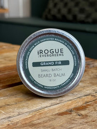 Close-up shows a tin of Rogue Evergreens Grand Fir beard balm made with natural ingredients on a wooden surface.
