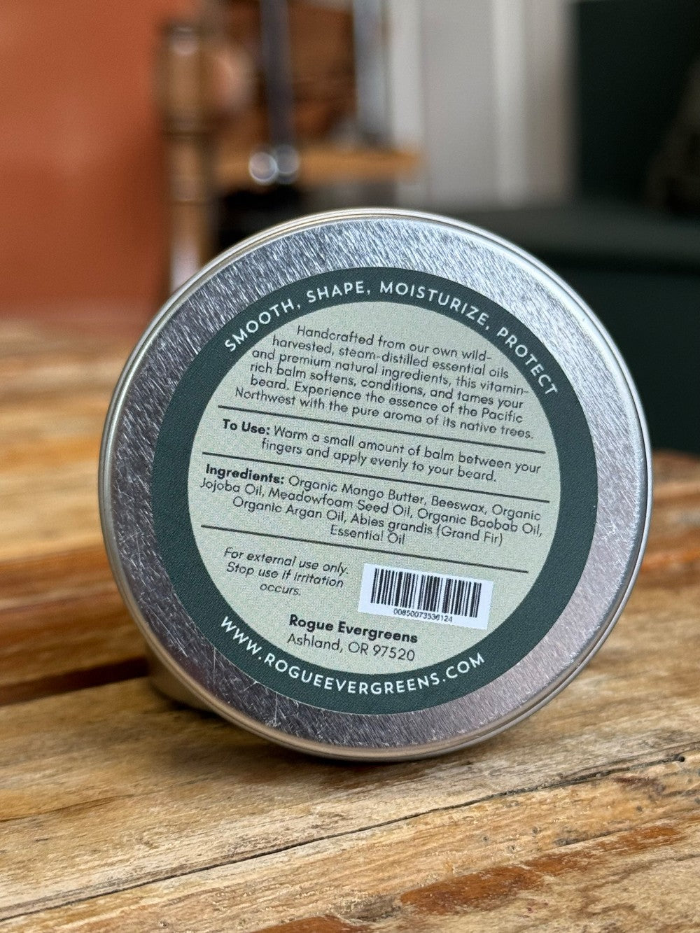 Close-up shows the label on a Rogue Evergreens beard balm tin, advertising natural ingredients like mango butter and fir essential oil to smooth, shape, moisturize, and protect beards.
