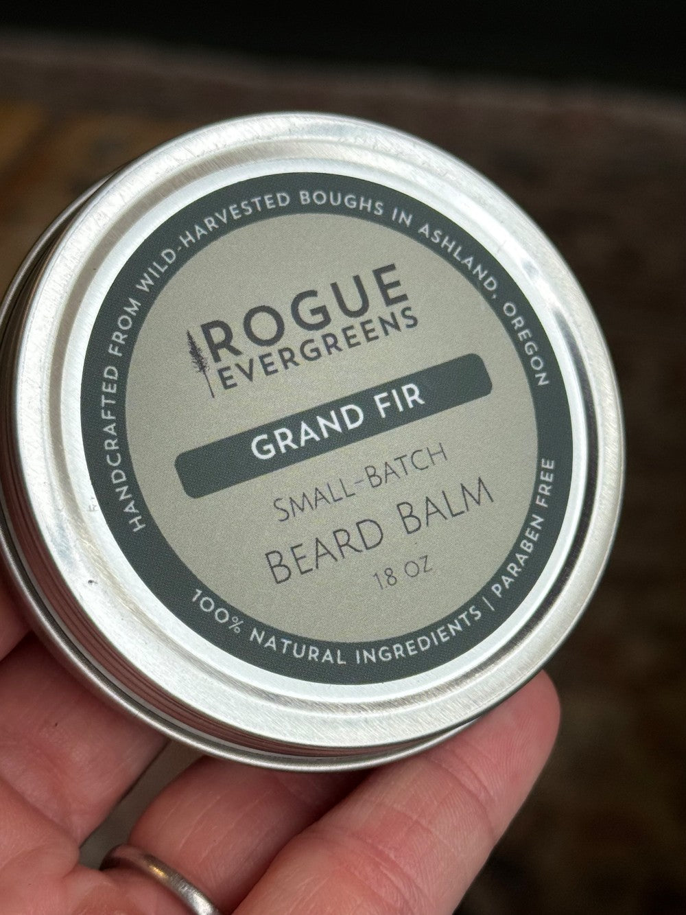 Hand holds a tin of Rogue Evergreens Grand Fir beard balm, showcasing its 100% natural ingredients.
