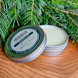 Rogue Evergreens beard balm, showcasing natural ingredients, sits on a wooden surface surrounded by fresh grand fir needles.
