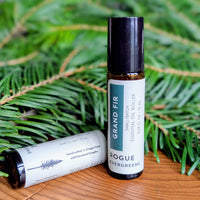 Two Rogue Evergreens Grand Fir essential oil roller bottles sit on a wooden surface amongst fir branches.