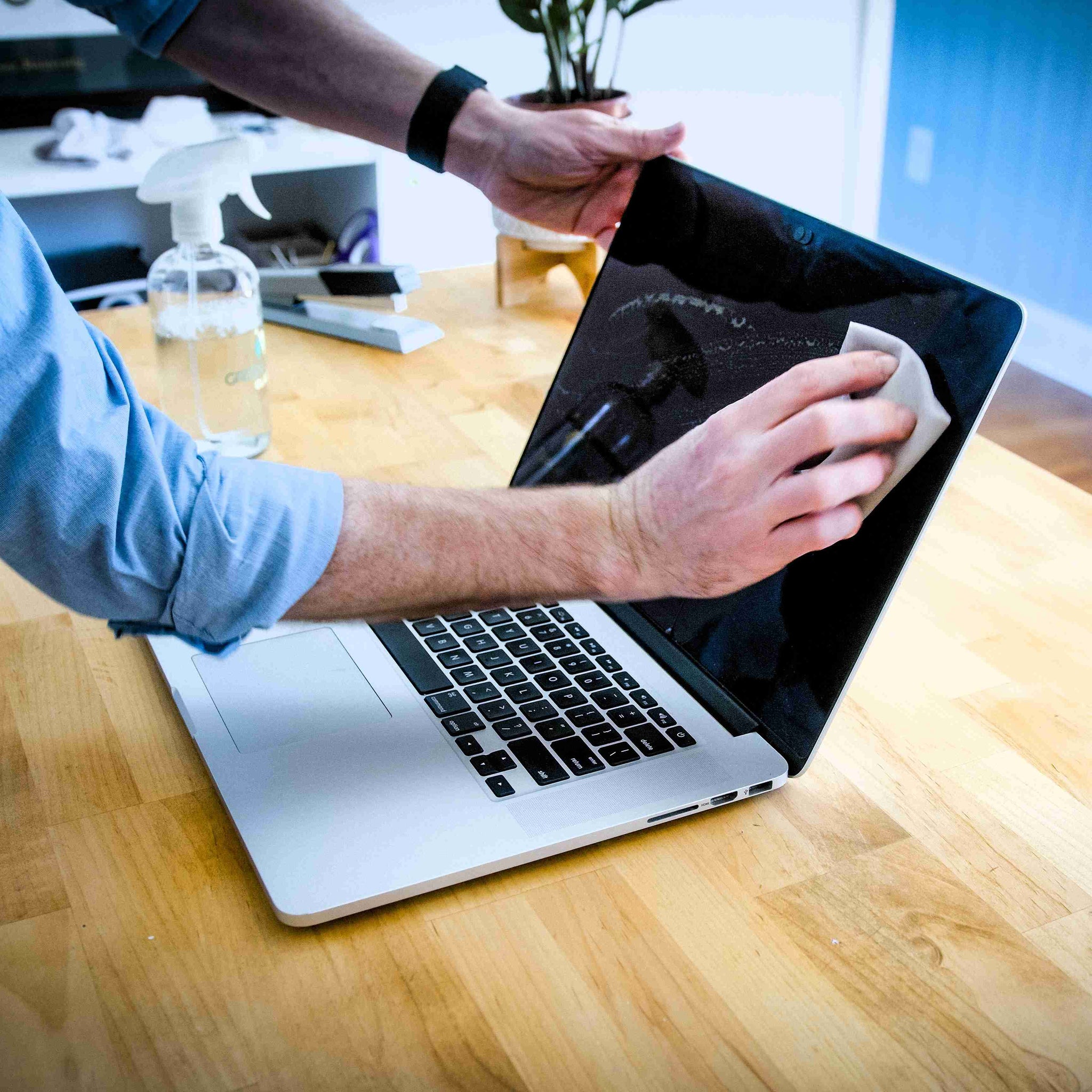 A person cleans the screen of a silver Apple laptop with a microfiber cloth and cleaning spray on a wooden table.
